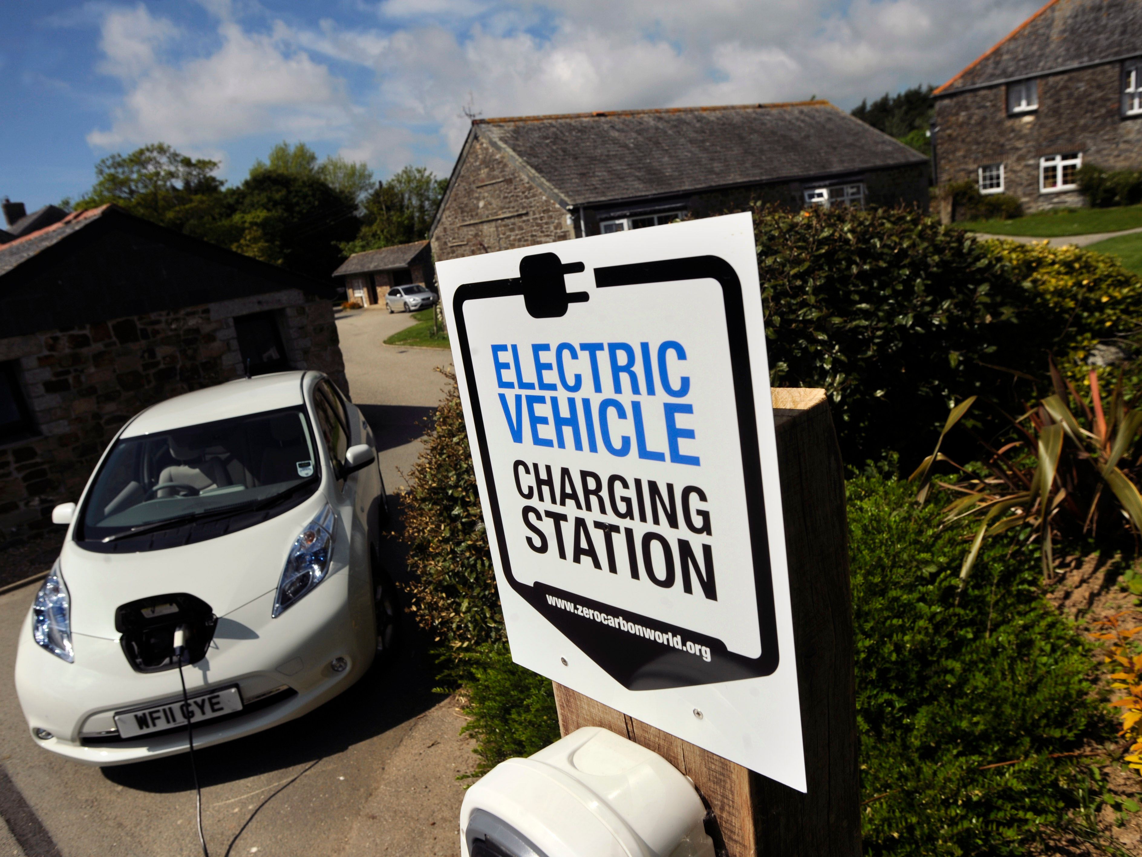 Electric vehicle charging station sign in front of a white electric car that is plugged in, with stone buildings and greenery in the background.