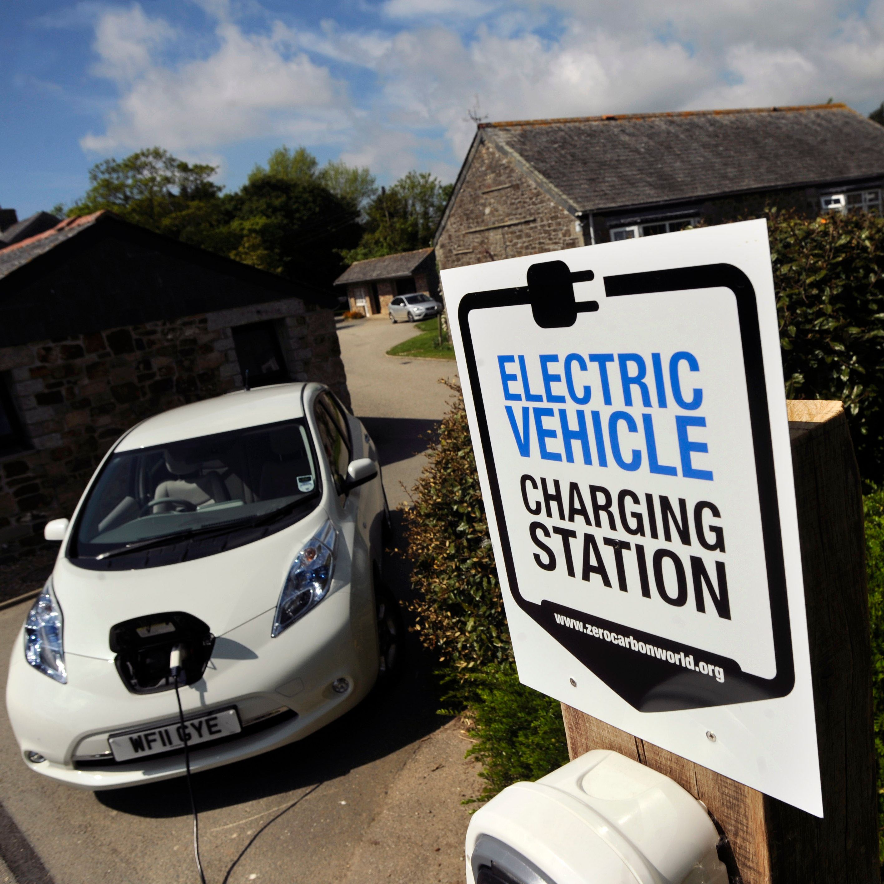 Electric vehicle charging station sign in front of a white electric car that is plugged in, with stone buildings and greenery in the background.