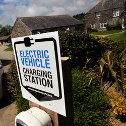 Electric vehicle charging station sign in front of a white electric car that is plugged in, with stone buildings and greenery in the background.