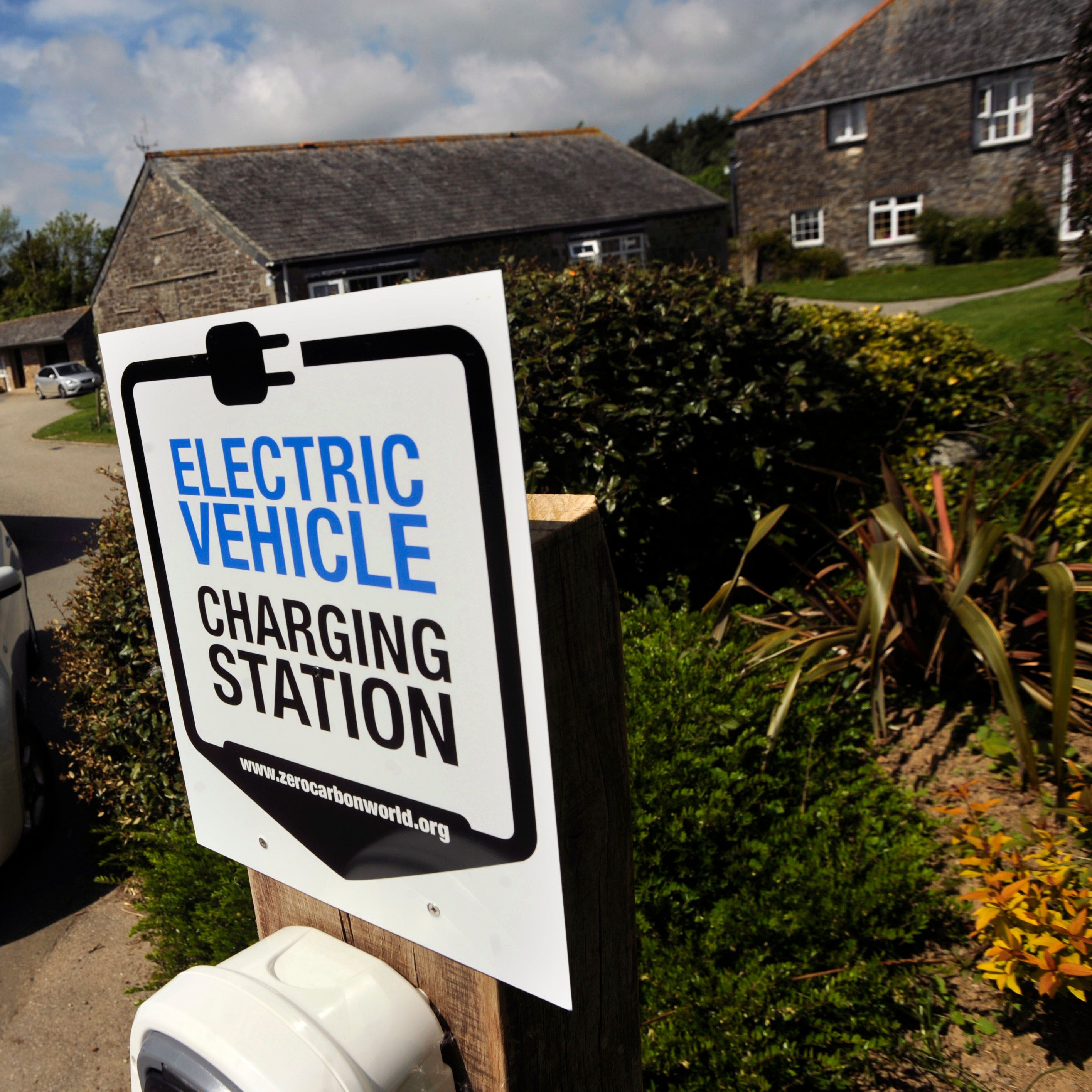 Electric vehicle charging station sign in front of a white electric car that is plugged in, with stone buildings and greenery in the background.