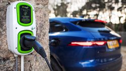 An electric vehicle charging at a wall-mounted EV charging station, with a blue car plugged in and trees in the background.
