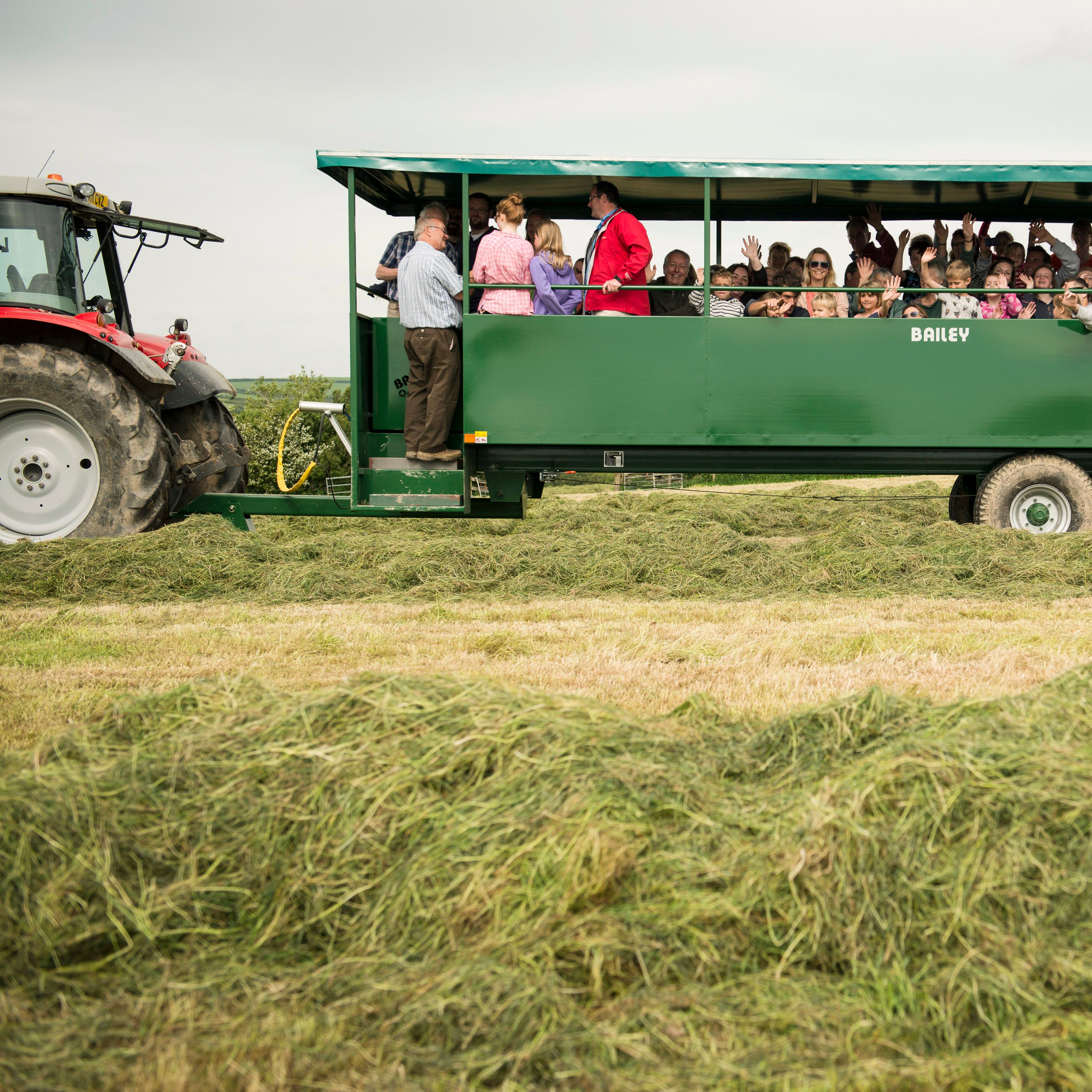 A group of people, including children, ride in a green wagon pulled by a tractor across a grassy field.