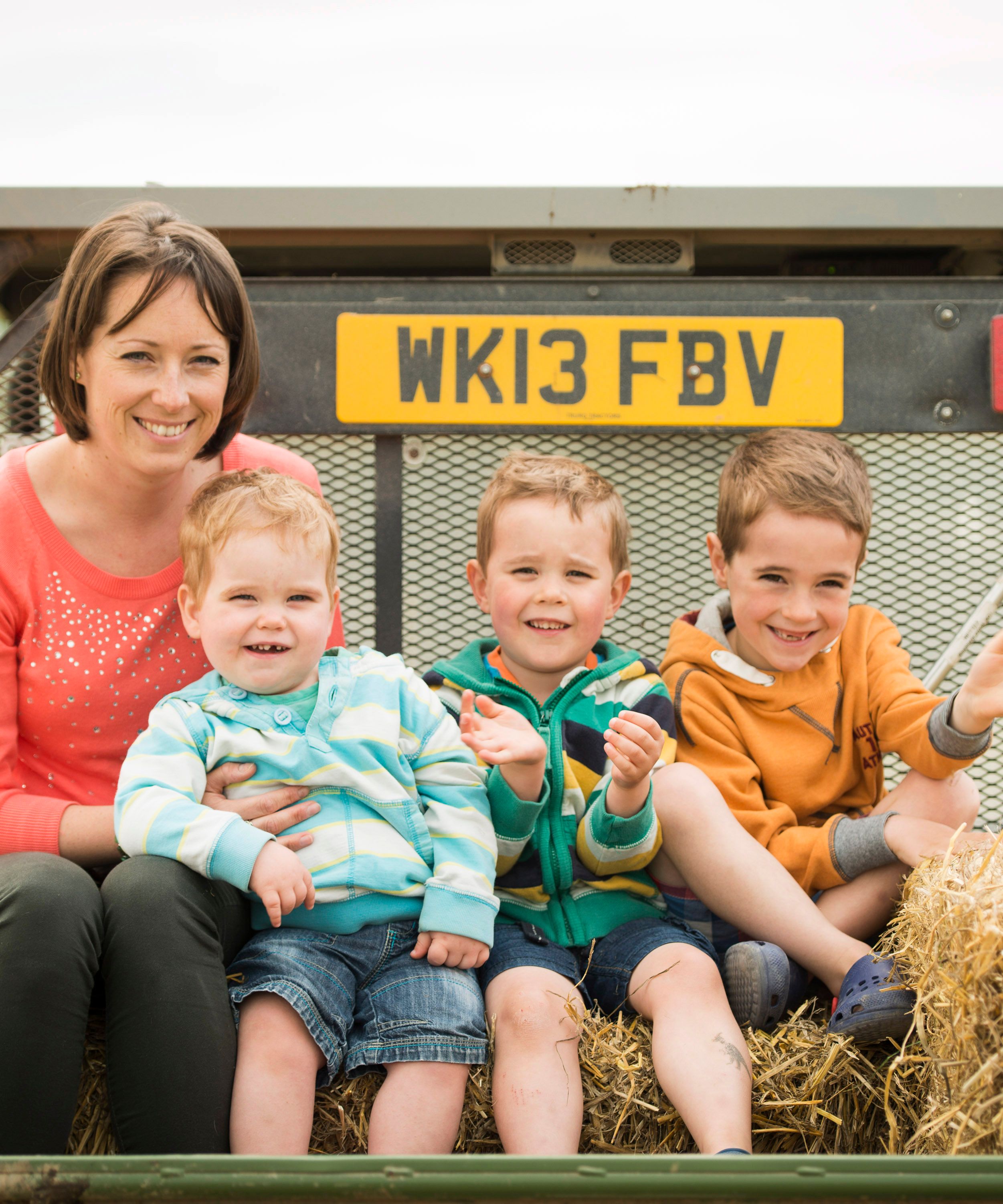 A woman and three young boys sitting on hay bales in the back of a trailer, smiling and enjoying an outdoor setting.