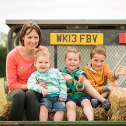 A woman and three young boys sitting on hay bales in the back of a trailer, smiling and enjoying an outdoor setting.