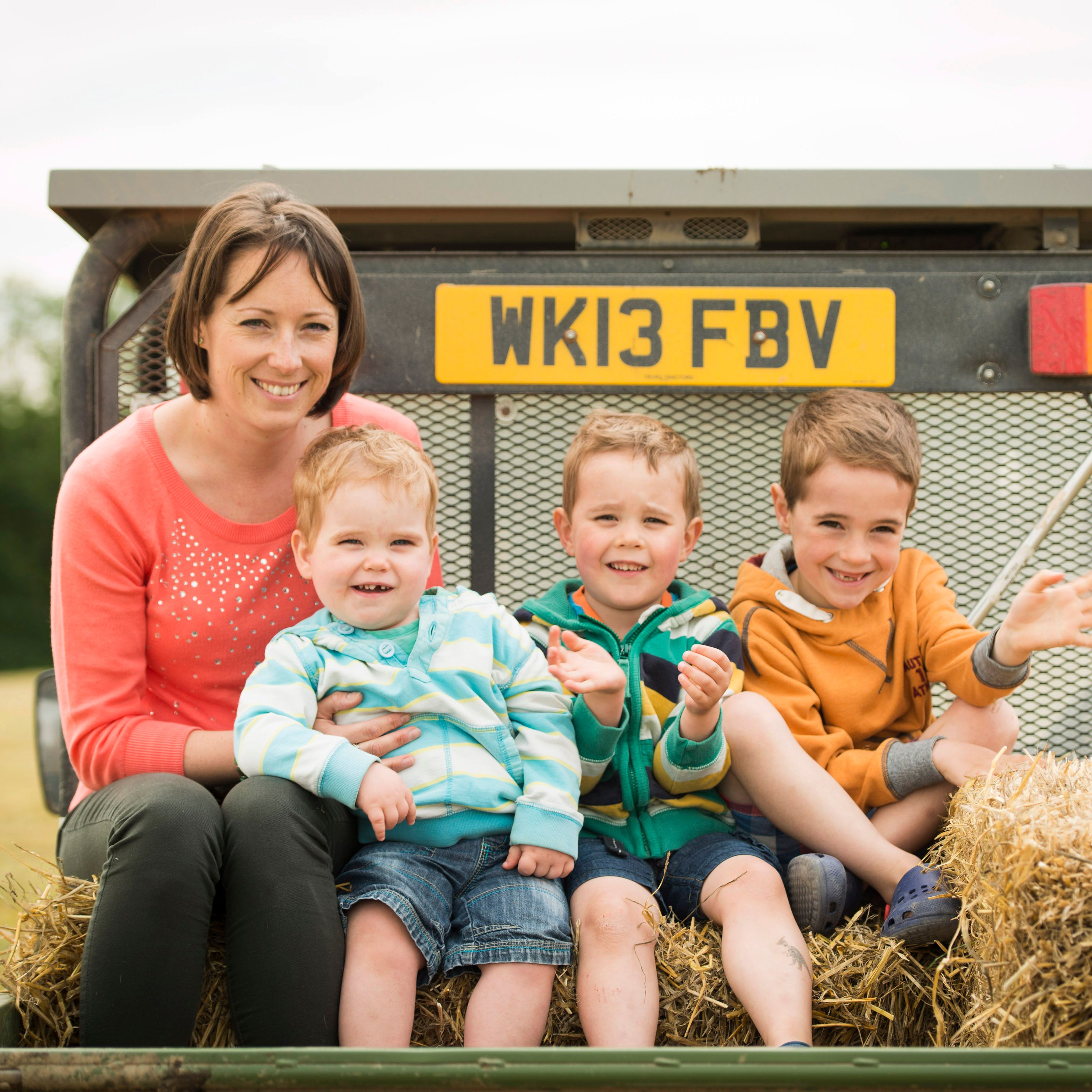 A woman and three young boys sitting on hay bales in the back of a trailer, smiling and enjoying an outdoor setting.