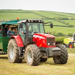 A red tractor pulling a green trailer filled with people, mostly children, on a farm with green rolling hills in the background. A yellow and white forage harvester is working in the field.