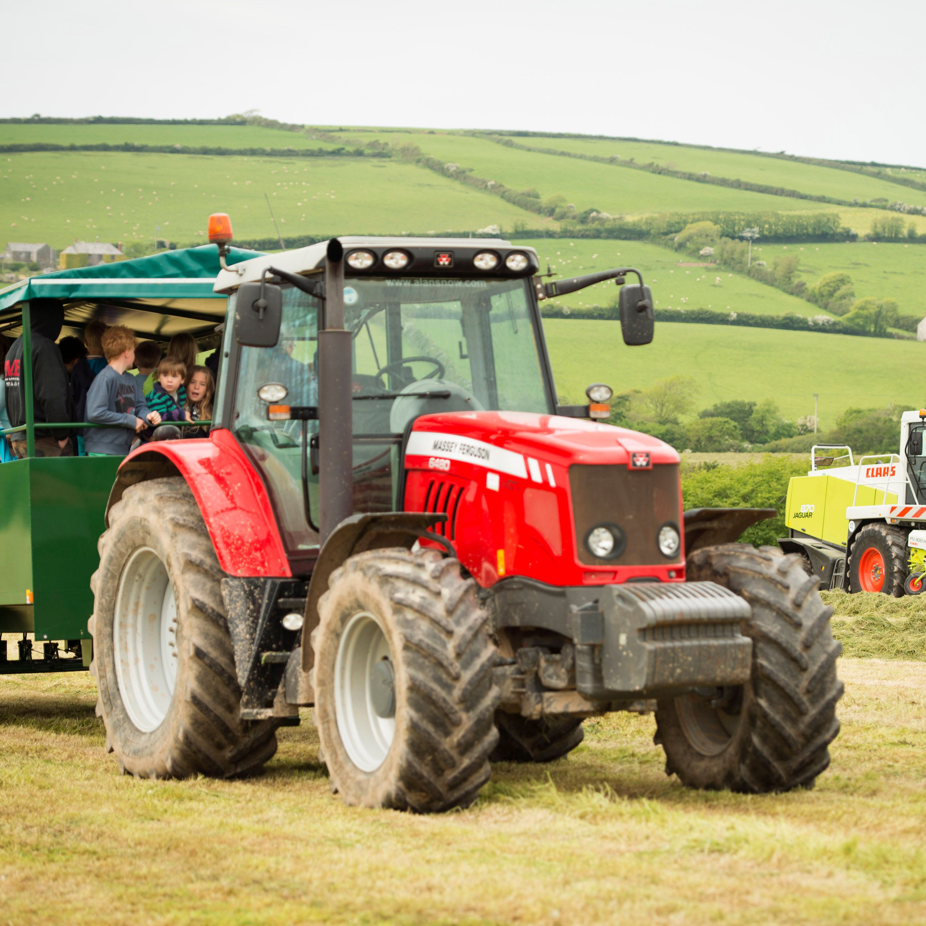A red tractor pulling a green trailer filled with people, mostly children, on a farm with green rolling hills in the background. A yellow and white forage harvester is working in the field.