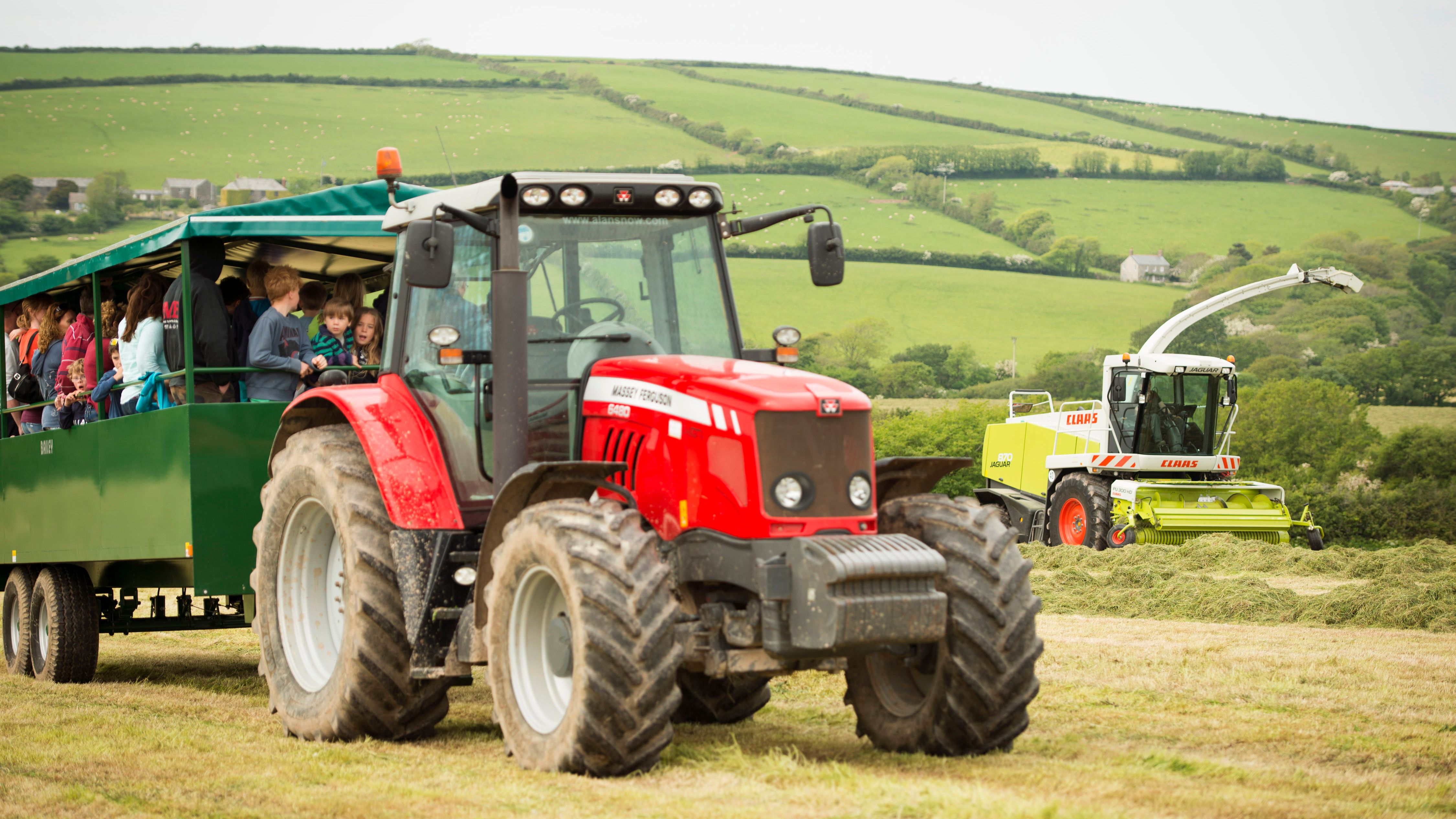 A red tractor pulling a green trailer filled with people, mostly children, on a farm with green rolling hills in the background. A yellow and white forage harvester is working in the field.