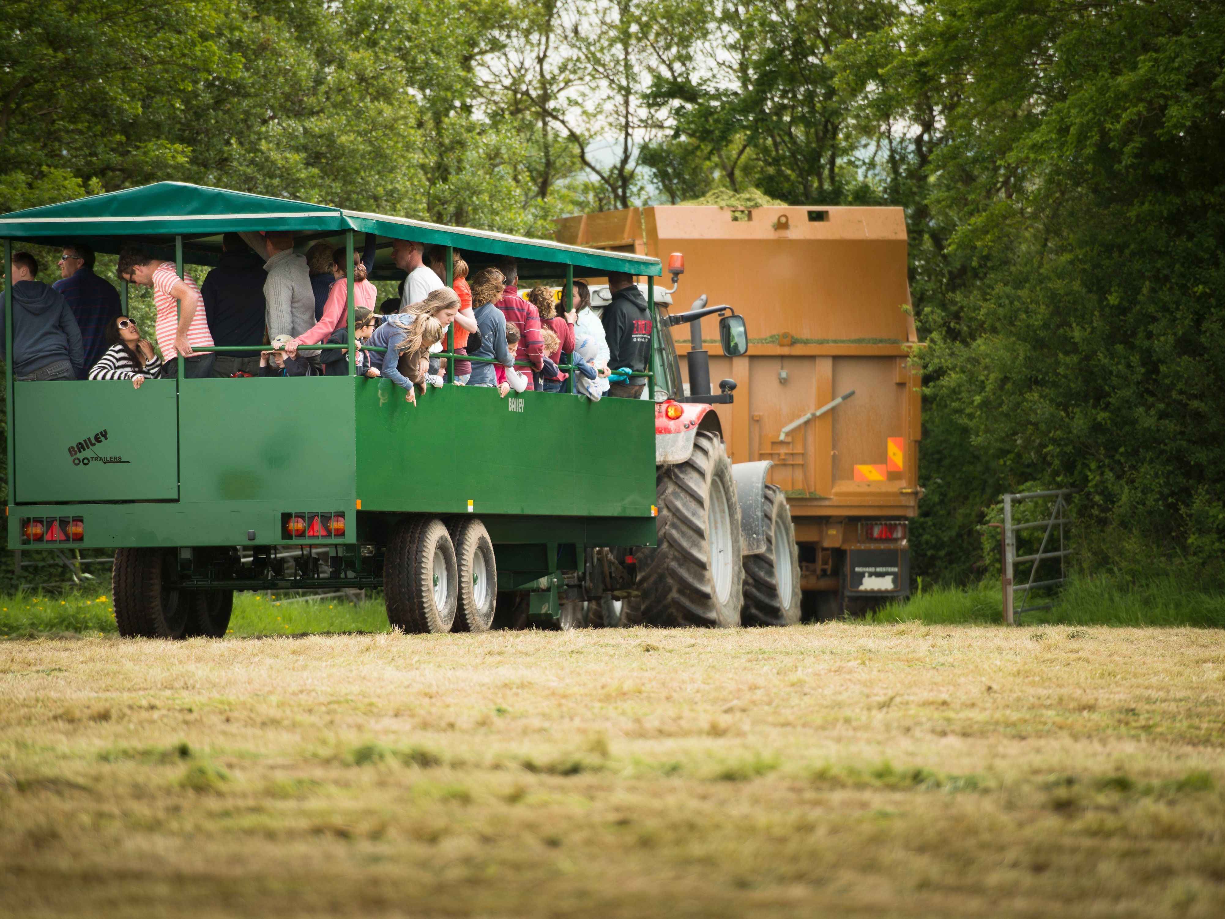 A green tractor pulling a covered trailer filled with people on a hayride through a grassy field, with trees and another farm vehicle in the background.