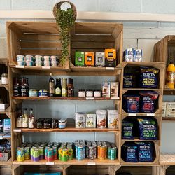 Wooden shelves displaying a variety of specialty groceries including snacks, canned drinks, sauces, pasta, and other packaged foods, with a hanging plant and branded reusable bags.