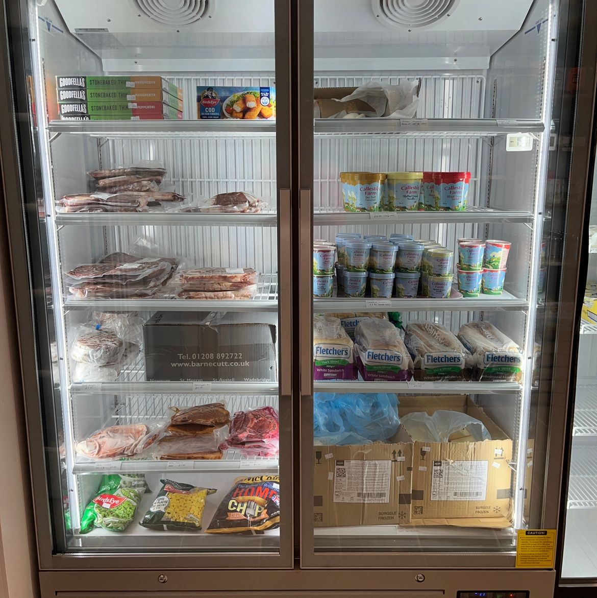 A commercial refrigerator with glass doors containing various frozen and refrigerated foods, including pizzas, packaged meats, vegetables, yogurts, bread, and boxes.