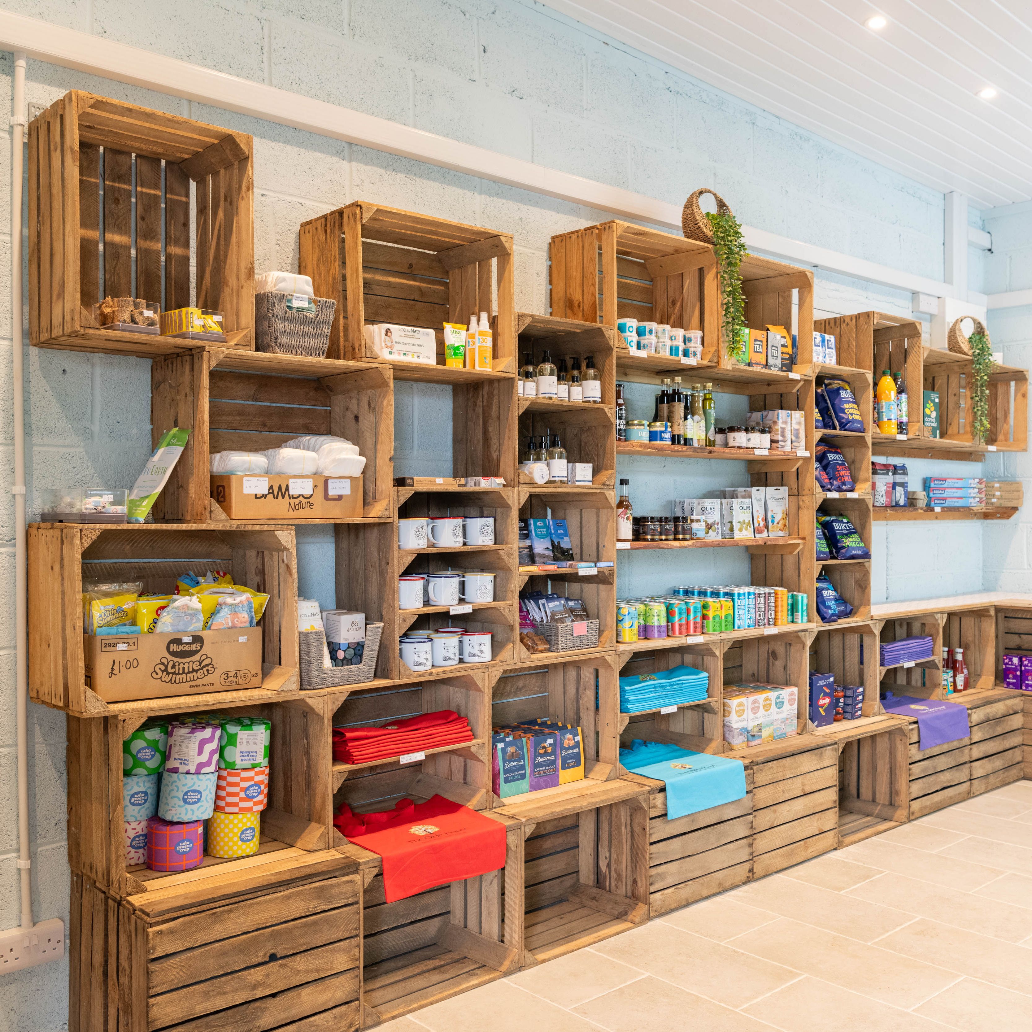 A small store with wooden crate shelves displaying a variety of products including snacks, drinks, mugs, household items, and merchandise in a light and neatly organized room.