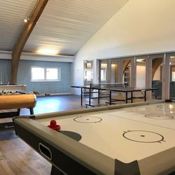 Spacious game room with an air hockey table, foosball table, and ping pong table, featuring wooden floors, brick walls, and glass doors.