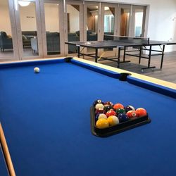 A game room featuring a blue pool table with racked billiard balls and a cue ball, and a ping pong table in the background. There is seating behind glass doors in an adjacent room.