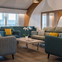 Modern living room with teal sofas, yellow and beige cushions, a wooden coffee table with a potted orchid, and large windows with light streaming in.