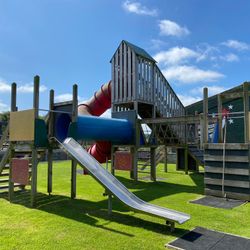 Outdoor playground structure with slides, climbing features, and tunnels on a grassy area under a blue sky.