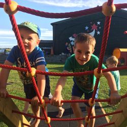 Two young boys climbing on a red rope playground structure outdoors with another child walking in the background, all enjoying a sunny day at a playground.