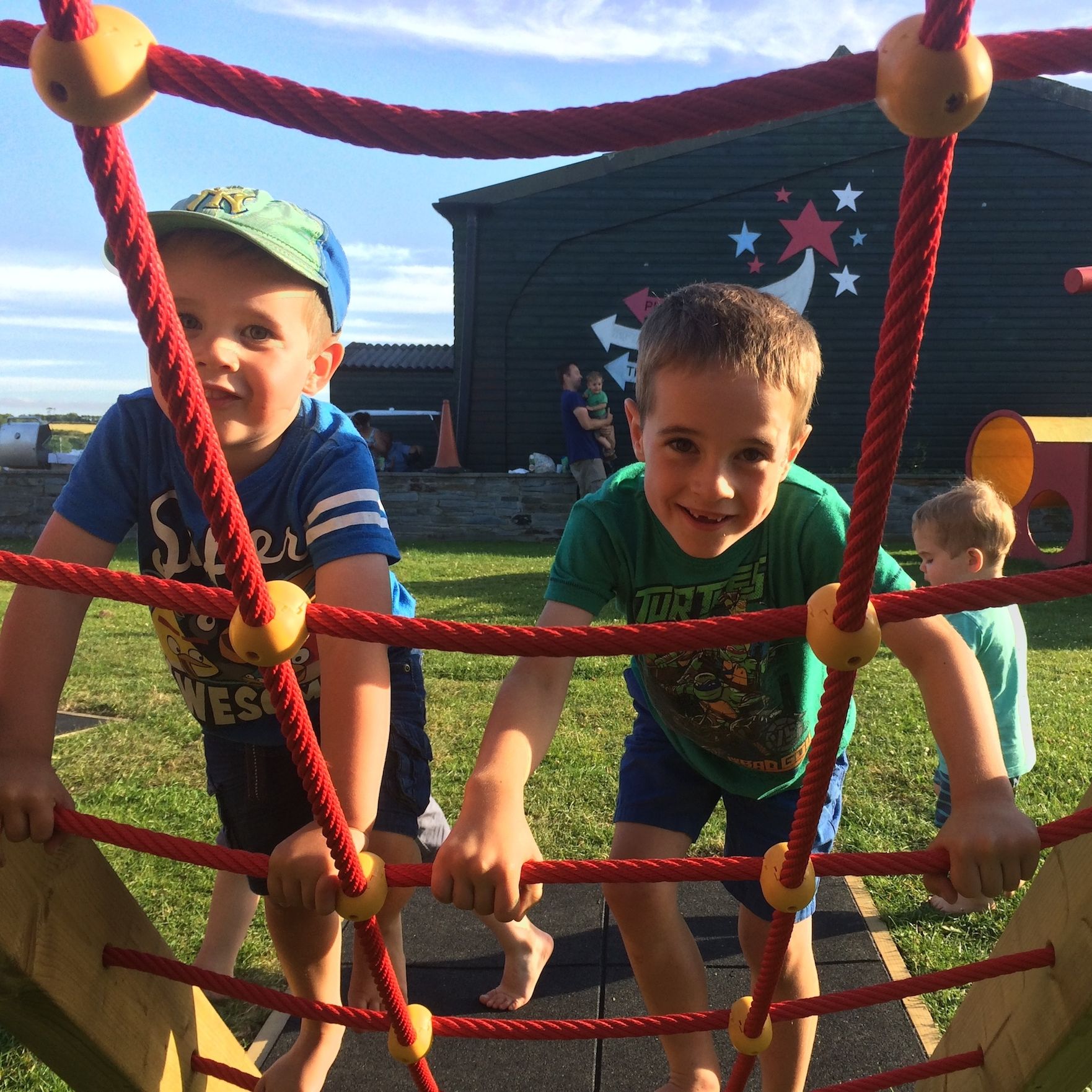 Two young boys climbing on a red rope playground structure outdoors with another child walking in the background, all enjoying a sunny day at a playground.