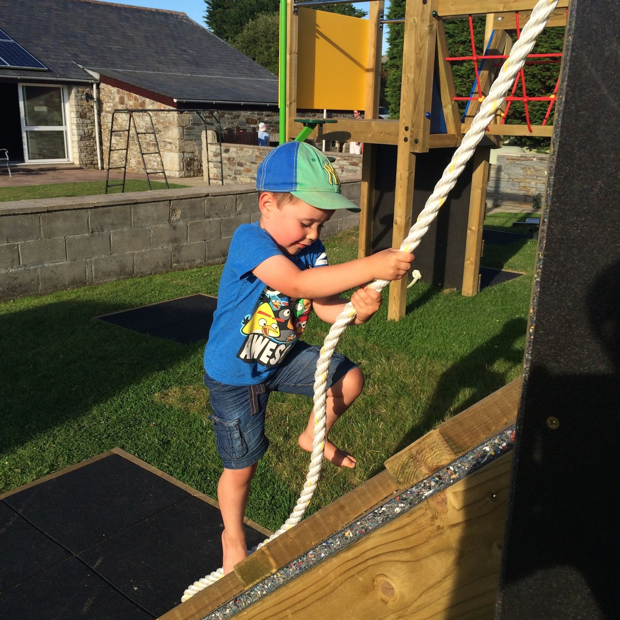 Young boy wearing a blue cap and t-shirt climbing up a wooden ramp with the help of a white rope at an outdoor playground on a sunny day.