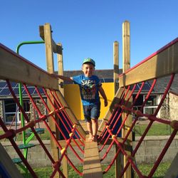 Young boy in a blue t-shirt and green cap walking barefoot on a wooden bridge structure with red ropes at a playground on a sunny day.