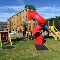 Children's playground with a wooden climbing structure, red tube slide, stairs, rope net, and grassy area under a blue sky.
