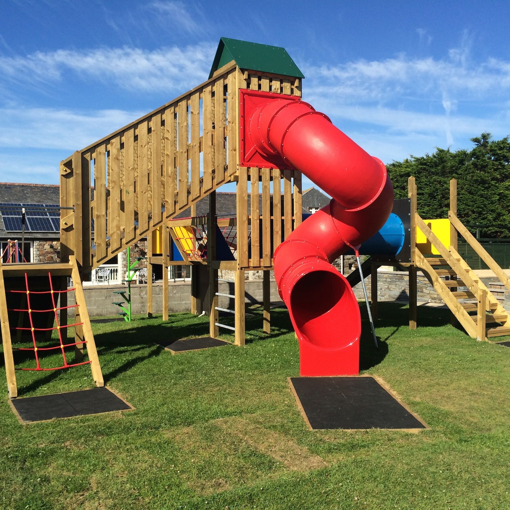 Children's playground with a wooden climbing structure, red tube slide, stairs, rope net, and grassy area under a blue sky.
