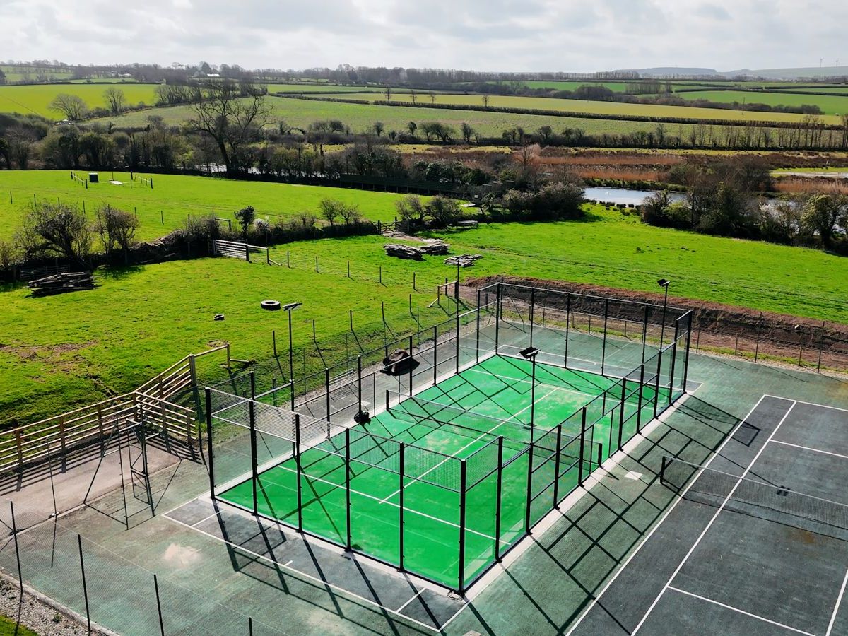 Aerial view of an outdoor tennis court surrounded by fences, set in a green rural landscape with fields, trees, and a river in the background.