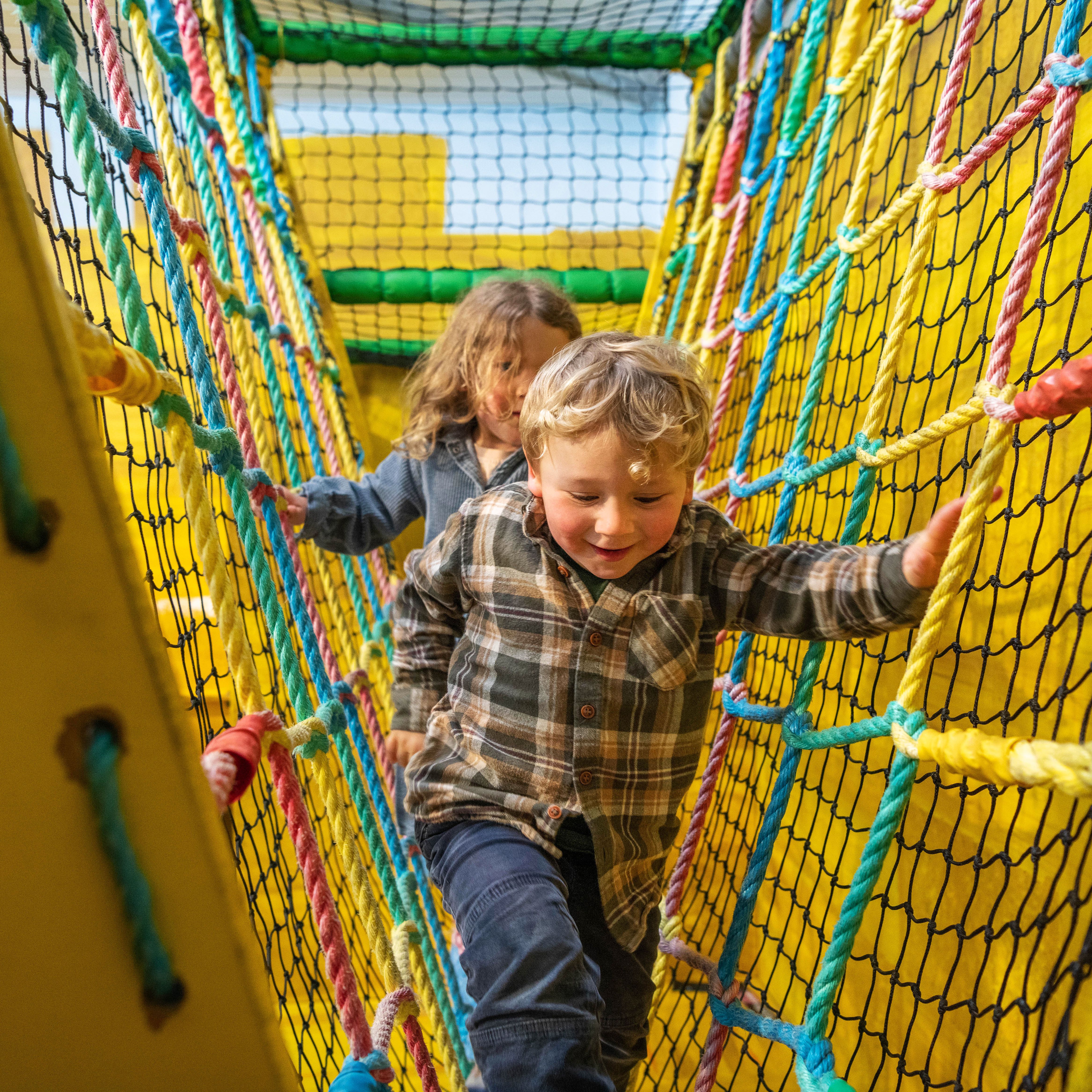 Two children climbing through a colorful rope bridge in an indoor play area with yellow padded walls.