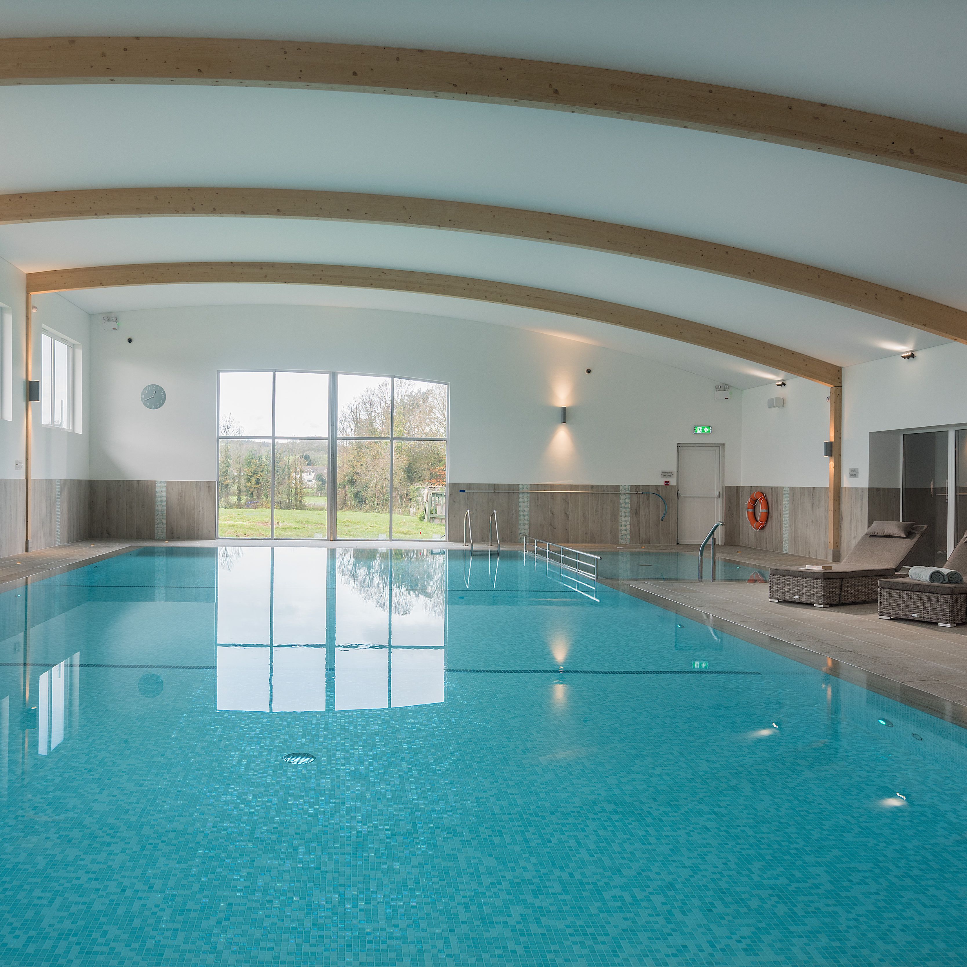 Modern indoor swimming pool with large windows, lounge chairs, and wooden beam ceiling.