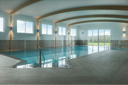 Modern indoor swimming pool with large windows, wooden beams on the ceiling, and natural light illuminating the space.