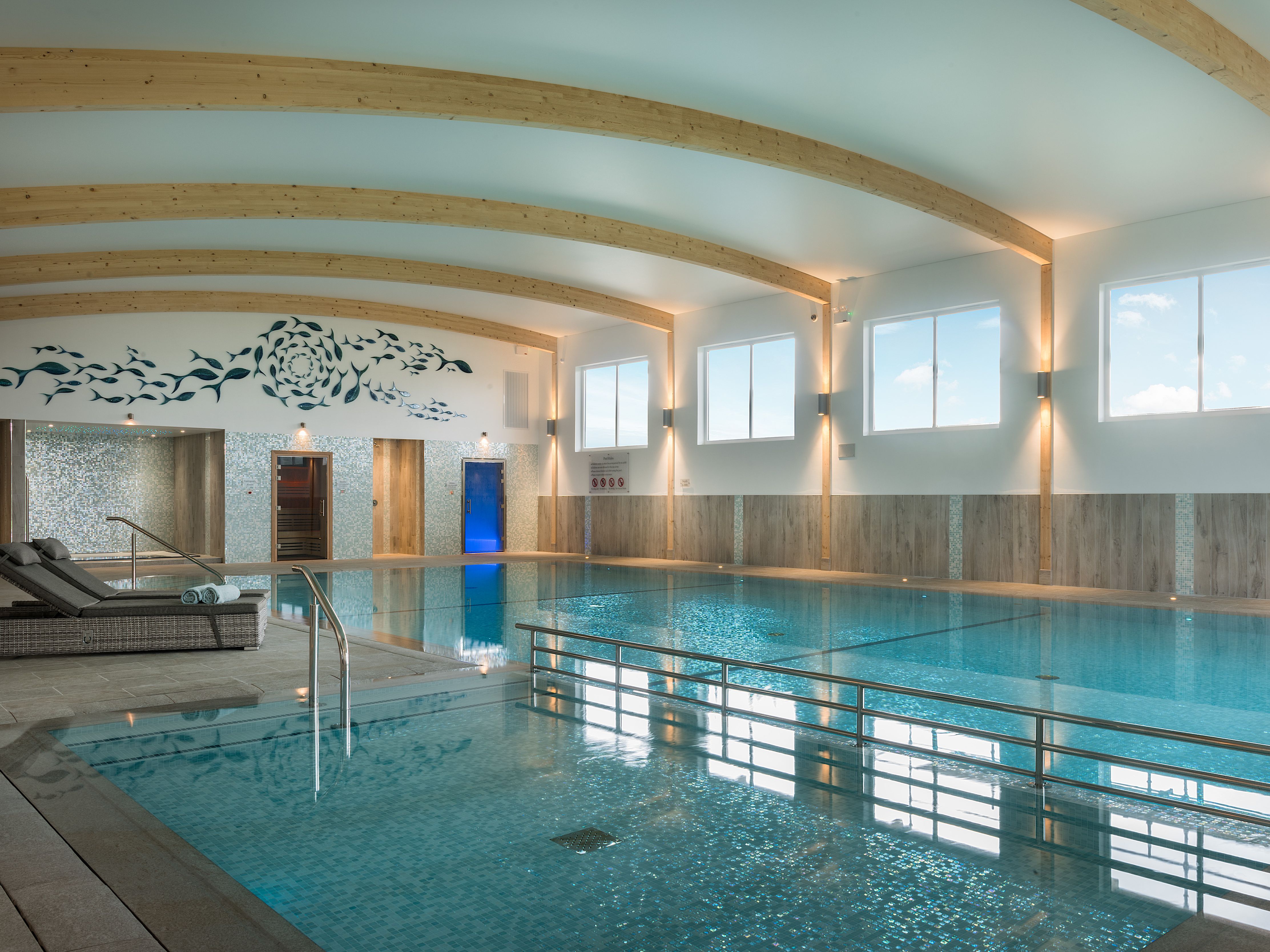 Modern indoor swimming pool with wooden beams, lounge chairs, and wall art featuring a fish design.