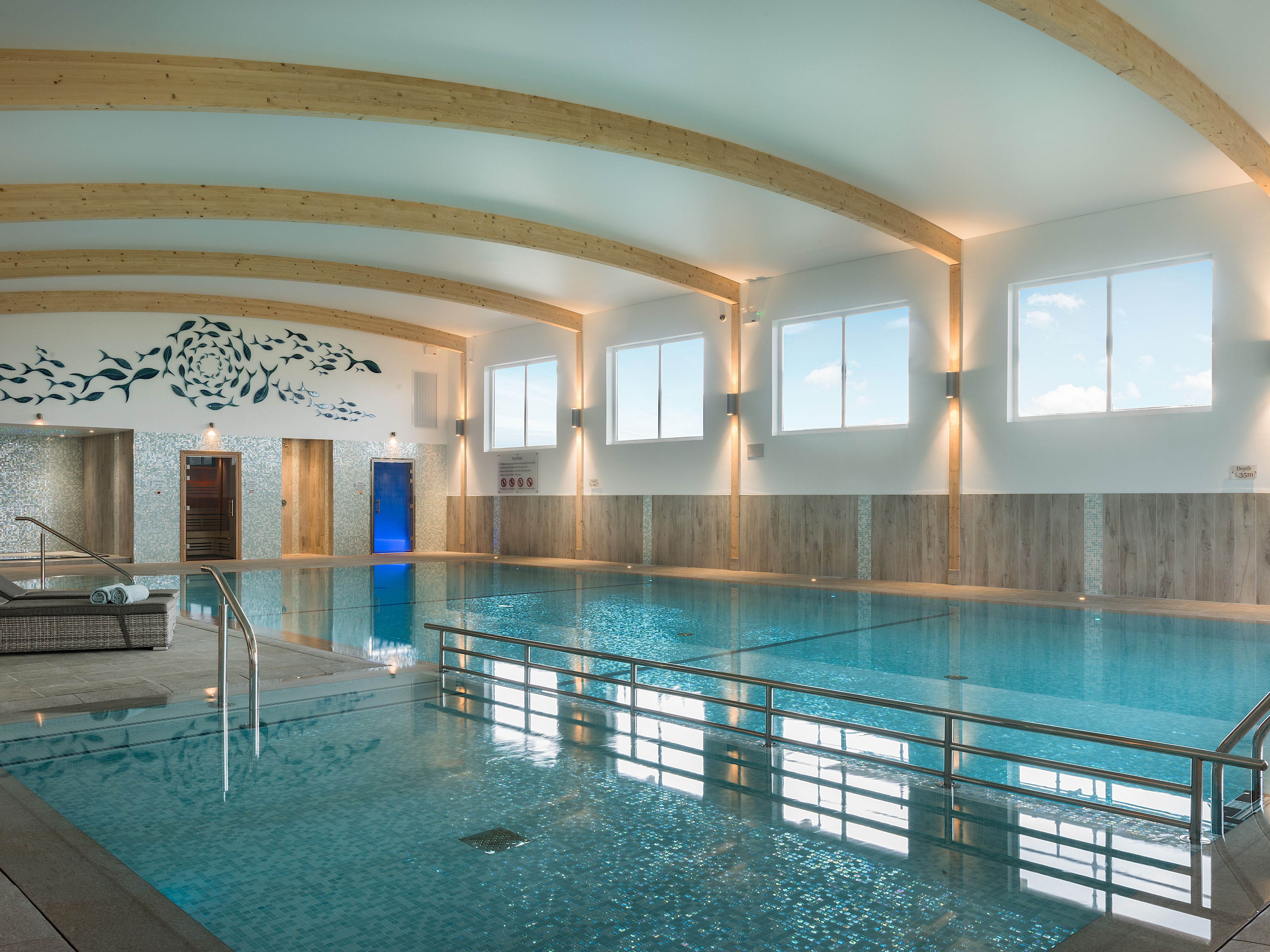 Modern indoor swimming pool with wooden beams, lounge chairs, and wall art featuring a fish design.