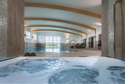 Modern indoor swimming pool area with wooden beam ceiling, a bubbling hot tub in the foreground, lounge chairs, and large windows letting in natural light.