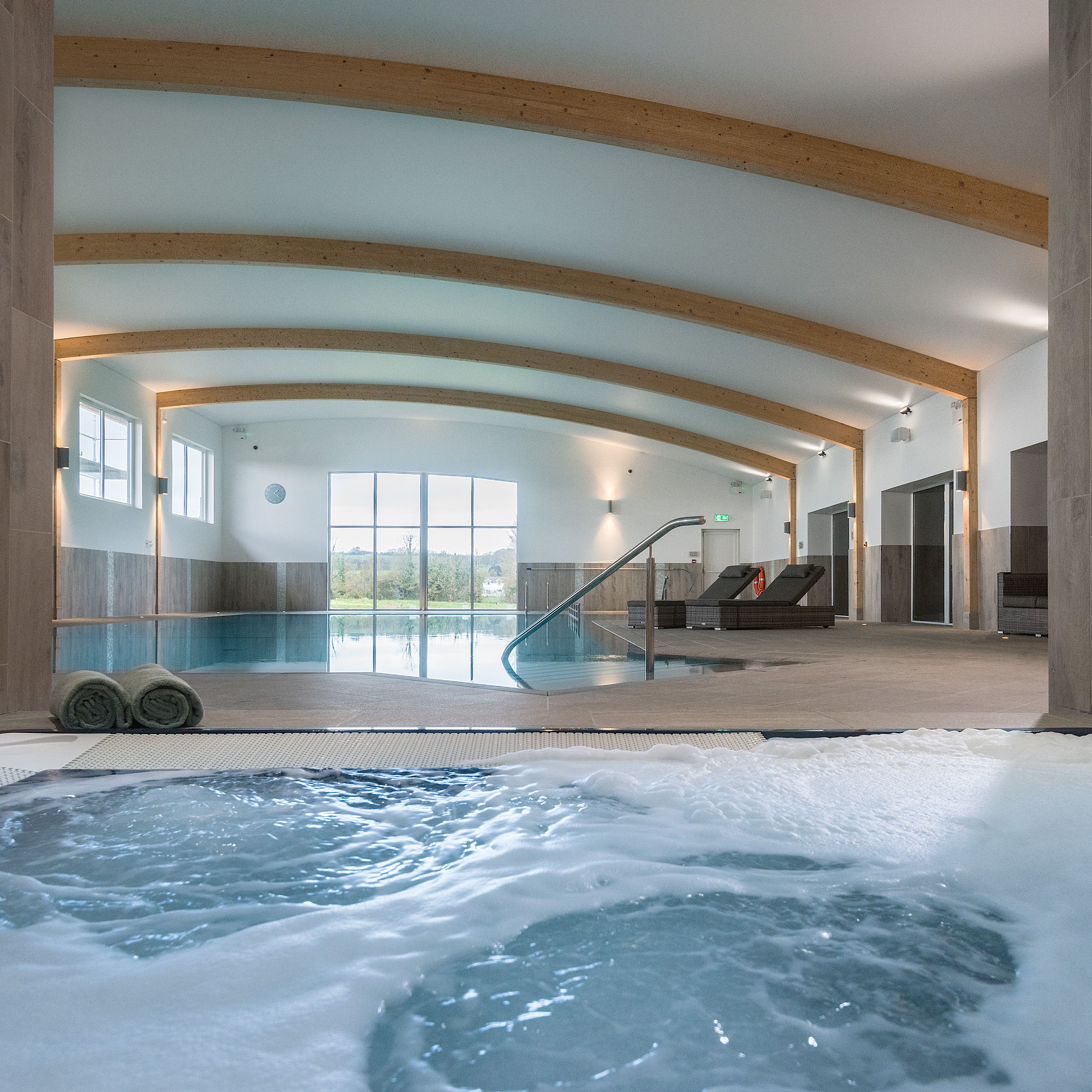 Modern indoor swimming pool area with wooden beam ceiling, a bubbling hot tub in the foreground, lounge chairs, and large windows letting in natural light.