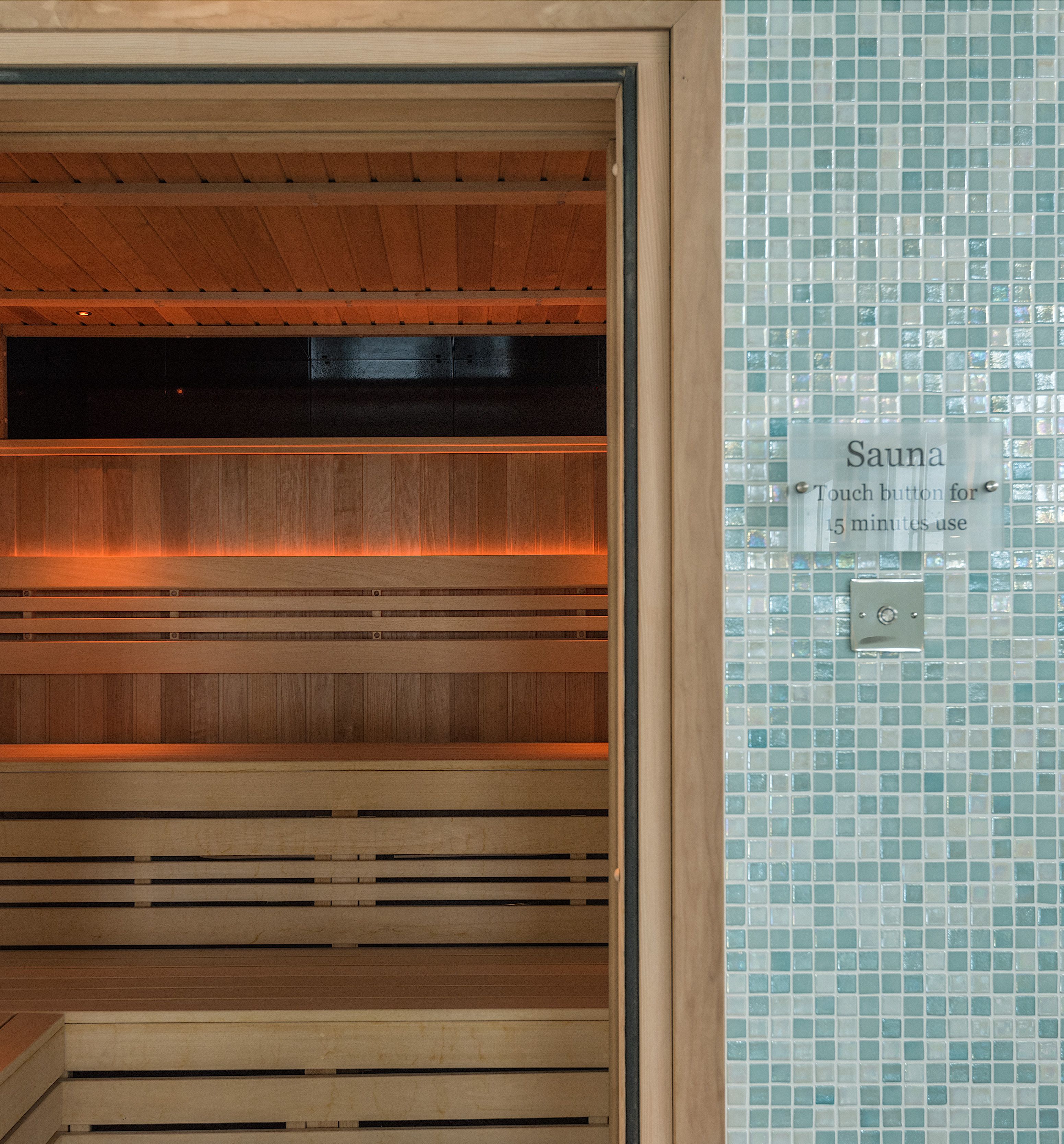 Modern wooden sauna with glass door, set in a room with light blue mosaic tile walls.