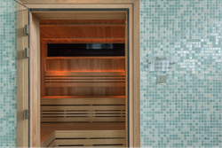 Modern wooden sauna with glass door, set in a room with light blue mosaic tile walls.