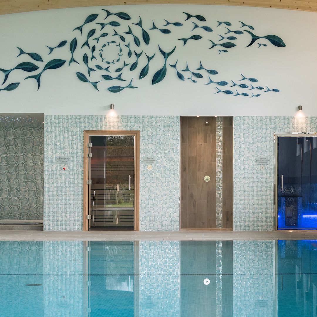 Indoor swimming pool area with mosaic tile walls, two glass doors, and a decorative fish art installation on the wall above. Natural light streams in through large windows on the right.