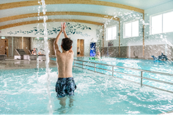 Boy playing in an indoor swimming pool with water splashing, other people relaxing and playing in the background.