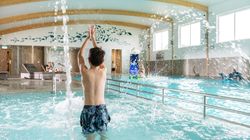 Boy playing in an indoor swimming pool with water splashing, other people relaxing and playing in the background.