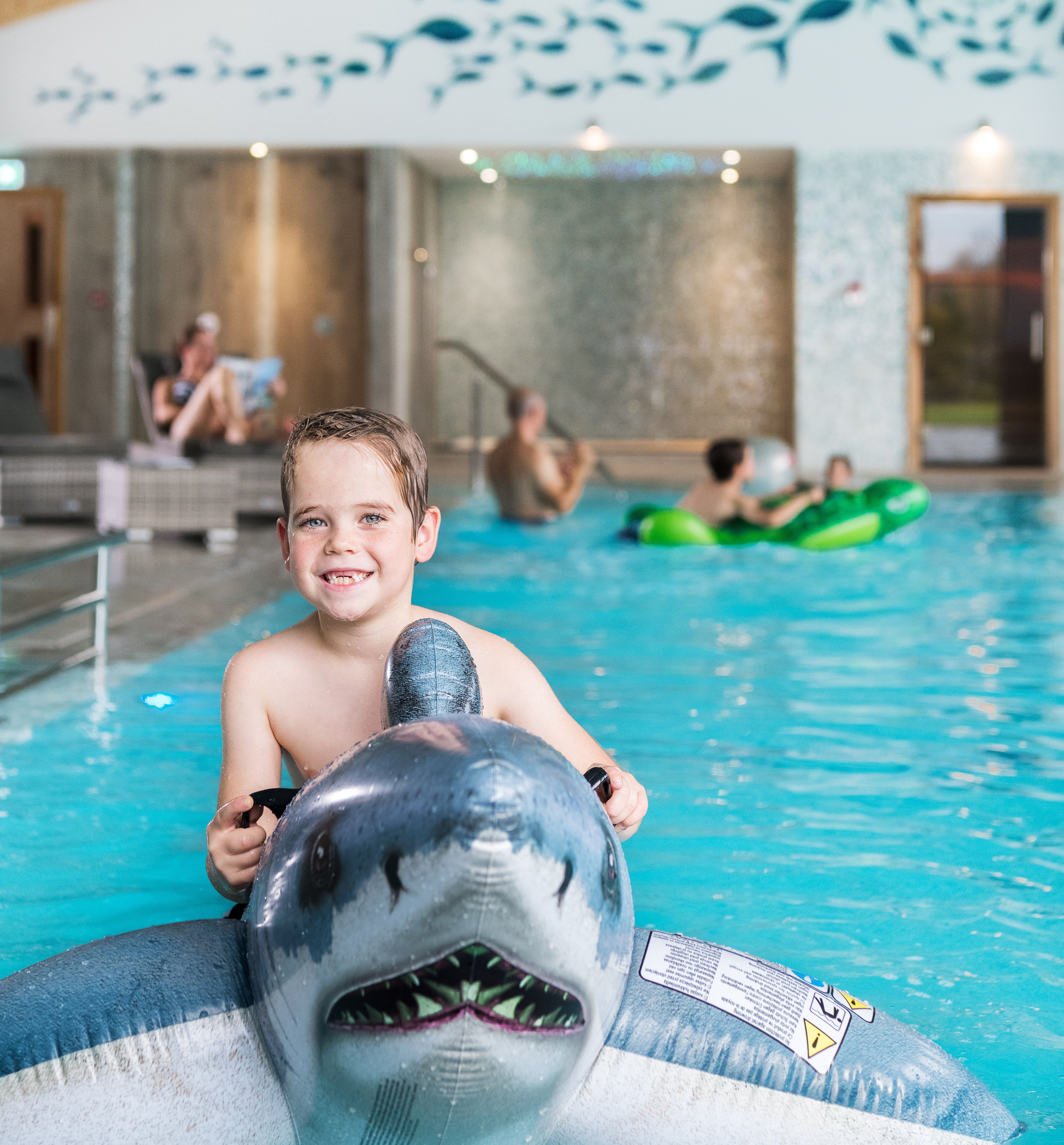 Smiling child riding an inflatable shark in an indoor swimming pool, with other people swimming and relaxing in the background.