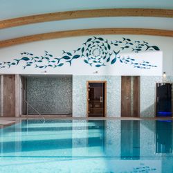 Indoor swimming pool with light blue tiles, wooden ceiling beams, mosaic walls, and a decorative fish mural above the pool area.