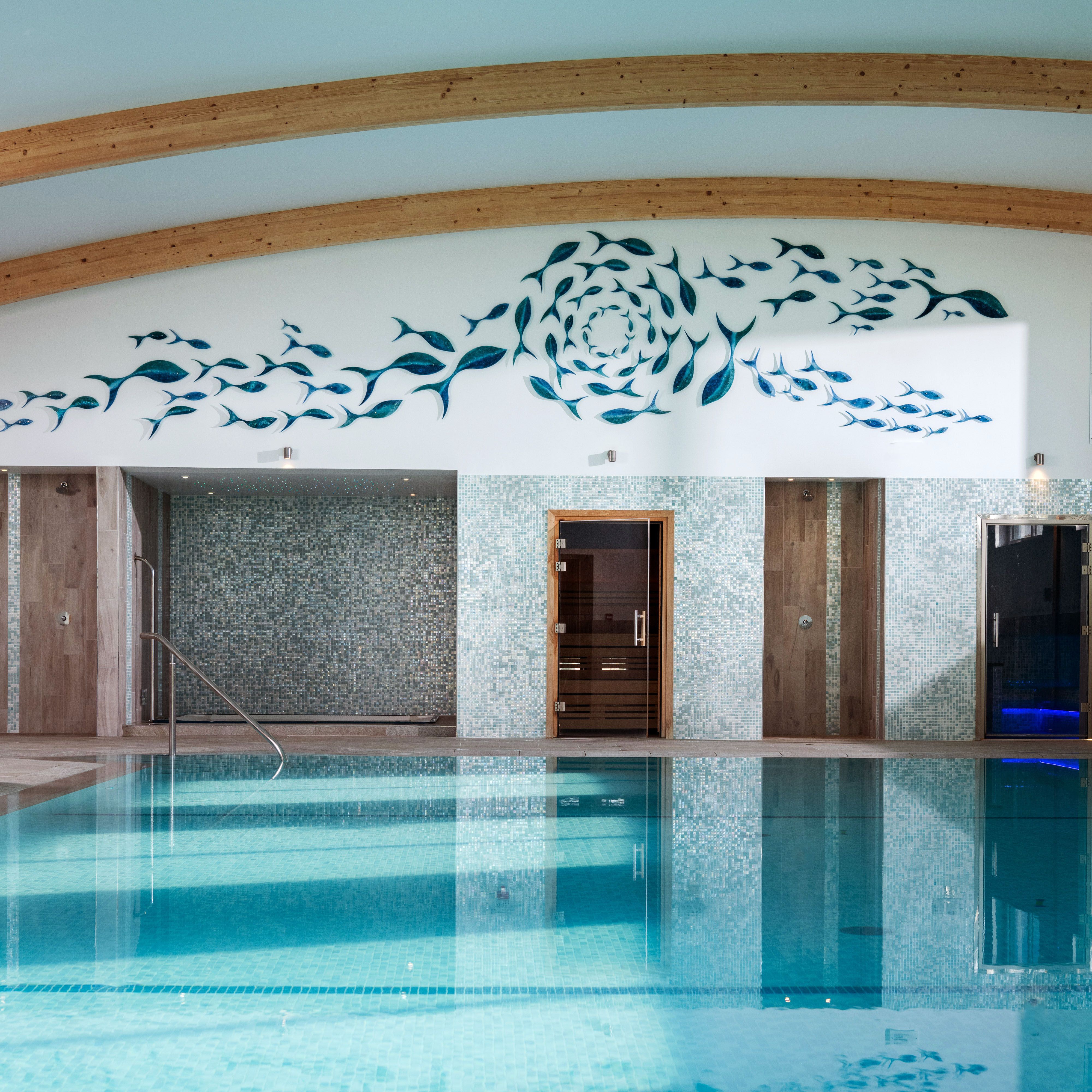 Indoor swimming pool with light blue tiles, wooden ceiling beams, mosaic walls, and a decorative fish mural above the pool area.