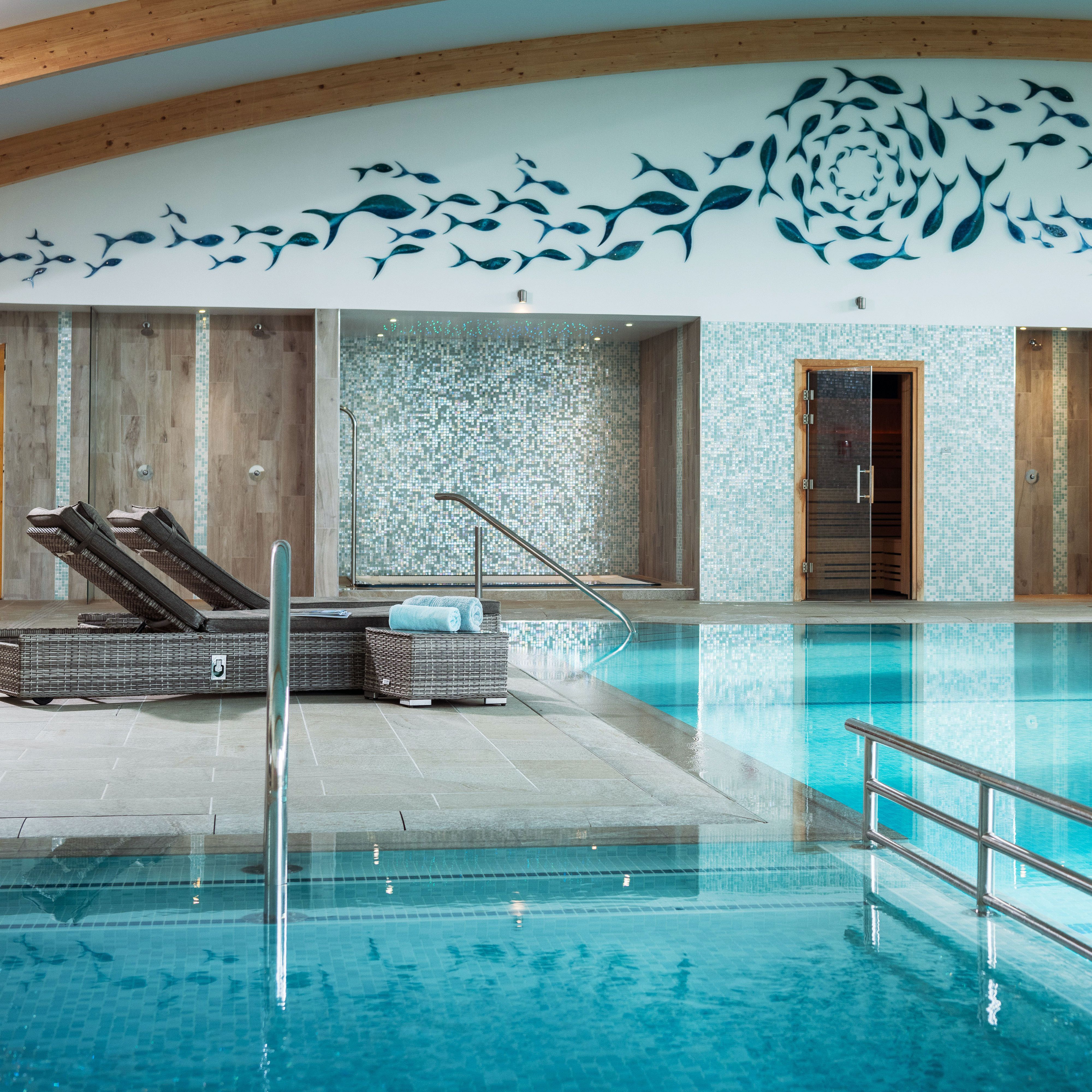 Modern indoor swimming pool with wicker lounge chairs, mosaic-tiled walls, sauna doors, turquoise water, and a decorative fish mural on the wall above.
