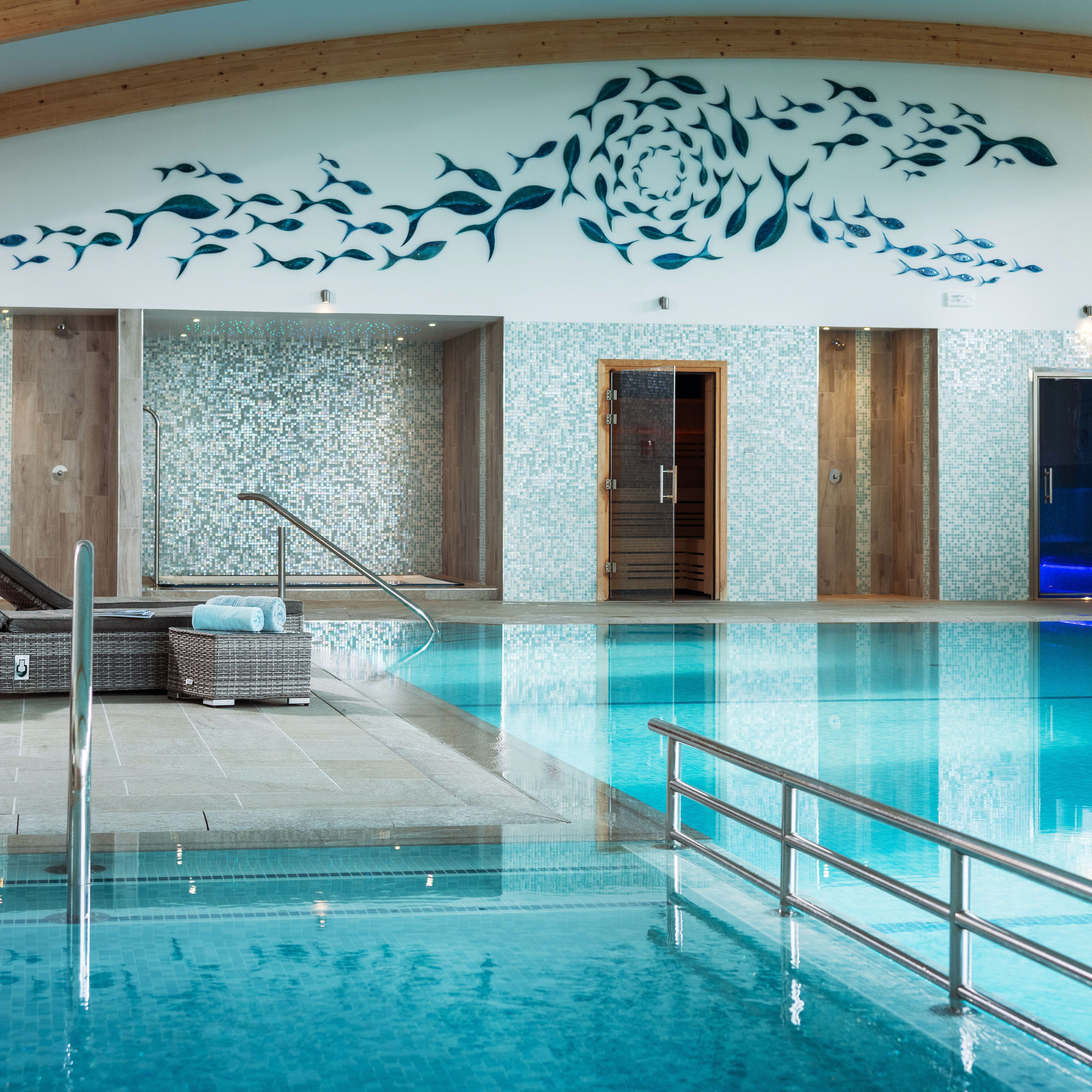 Modern indoor swimming pool with wicker lounge chairs, mosaic-tiled walls, sauna doors, turquoise water, and a decorative fish mural on the wall above.