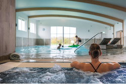 Woman relaxing in a hot tub at an indoor pool facility with people swimming and lounging in the background.