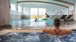 Woman relaxing in a hot tub at an indoor pool facility with people swimming and lounging in the background.