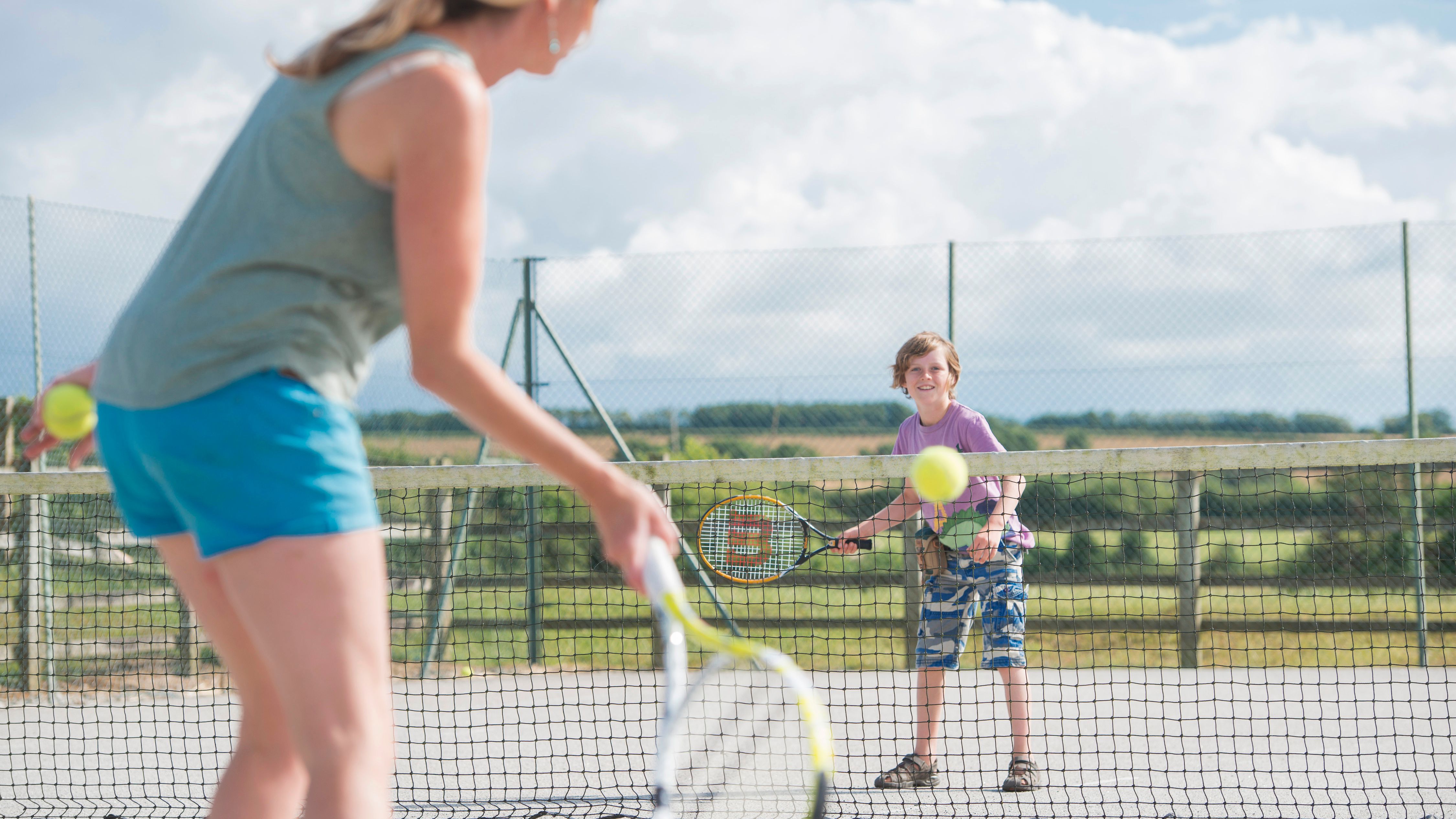 Woman and boy playing tennis on an outdoor court, with the woman in the foreground and the boy in the background, both holding rackets.