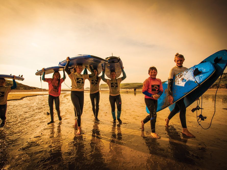 A group of children walking on the beach carrying surfboards, smiling and wearing wetsuits, with a warm sunset light in the background.