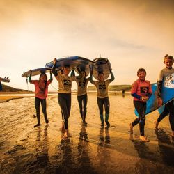 A group of children walking on the beach carrying surfboards, smiling and wearing wetsuits, with a warm sunset light in the background.