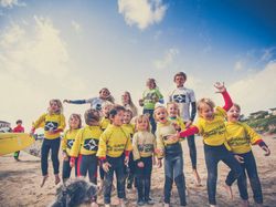 Group of children and instructors in wetsuits and yellow surf school shirts posing on a sandy beach, some jumping and smiling, with surfboards and a blue sky in the background.